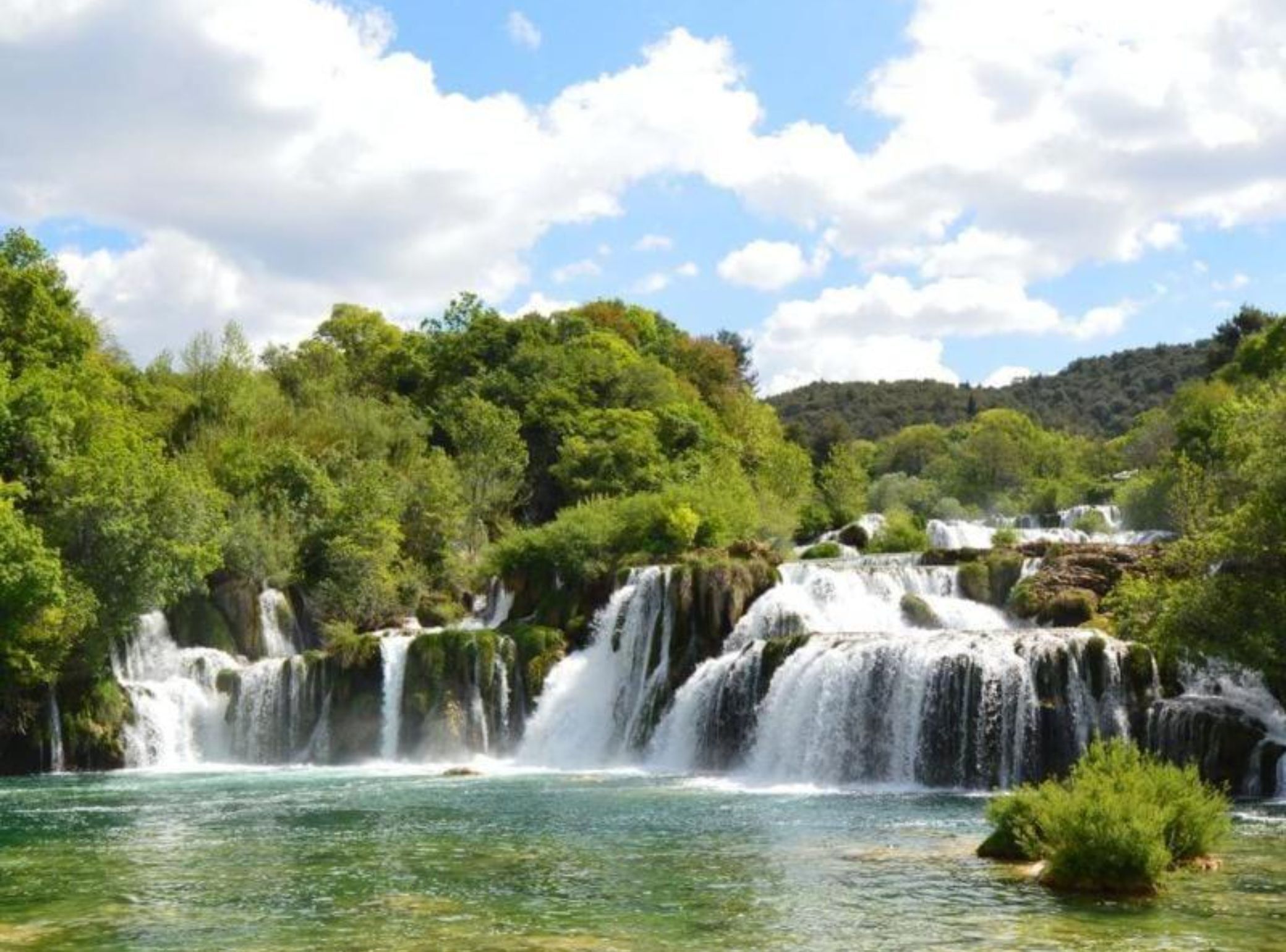 A waterfall cascading over rocks into a clear river surrounded by lush greenery and a forested hillside.
