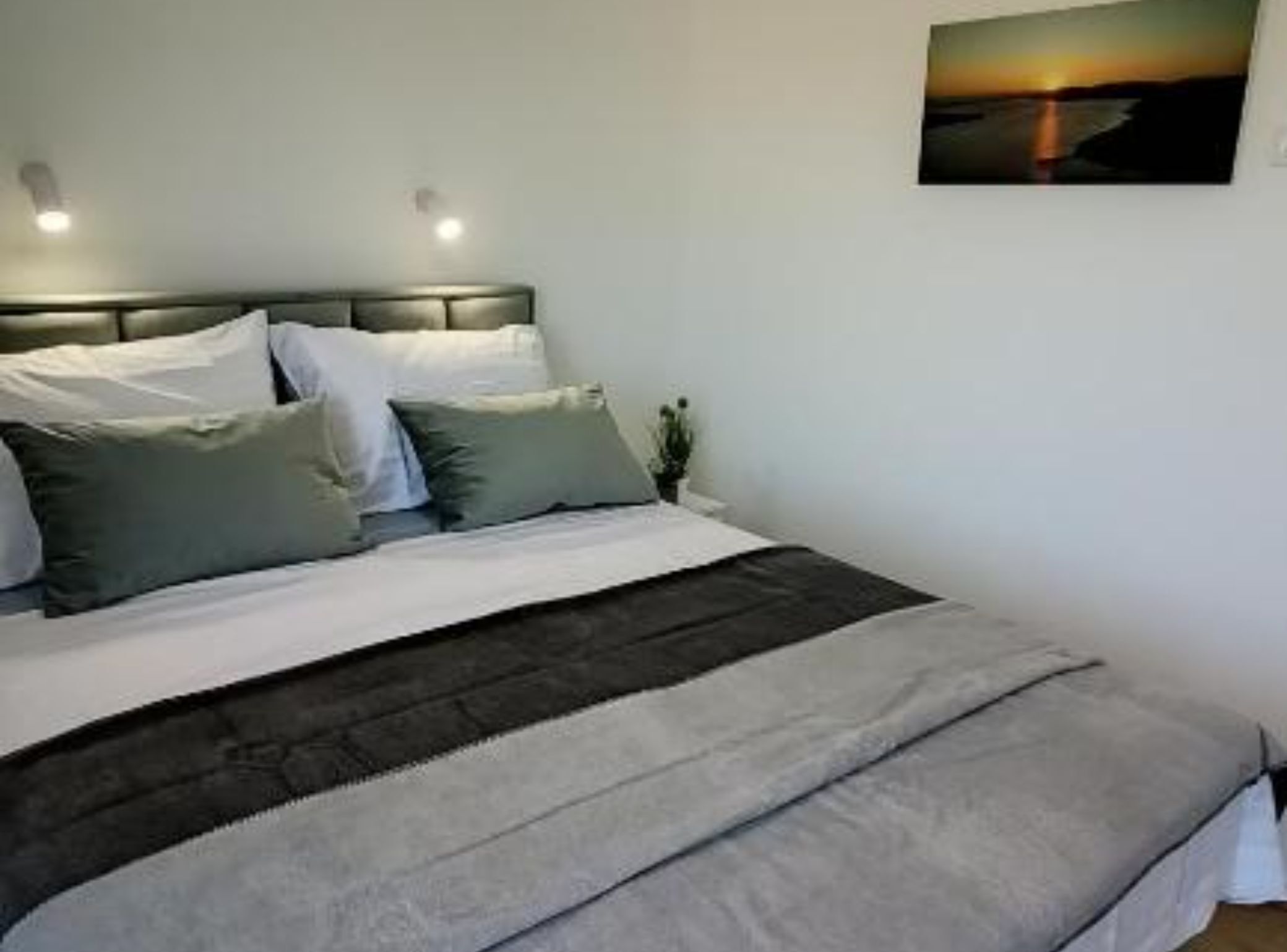 A neatly made bed with a grey and white striped blanket, a headboard with two wall-mounted lights, and a painting of a sunset on the wall.