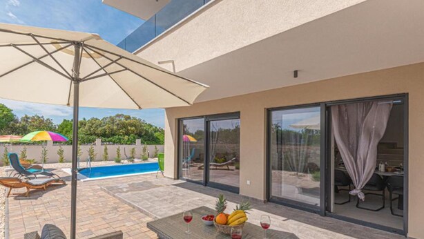 Une terrasse extérieure moderne, dotée d’une piscine, de chaises longues et d’une table avec des fruits et des verres à vin.