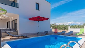 Un espace piscine moderne avec une piscine à l’eau d’un bleu limpide, des chaises longues et un parasol rouge.