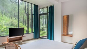 Une chambre avec une grande fenêtre, un téléviseur à écran plat, un banc en bois, un petit sac et un lit avec une tête de lit bleue.