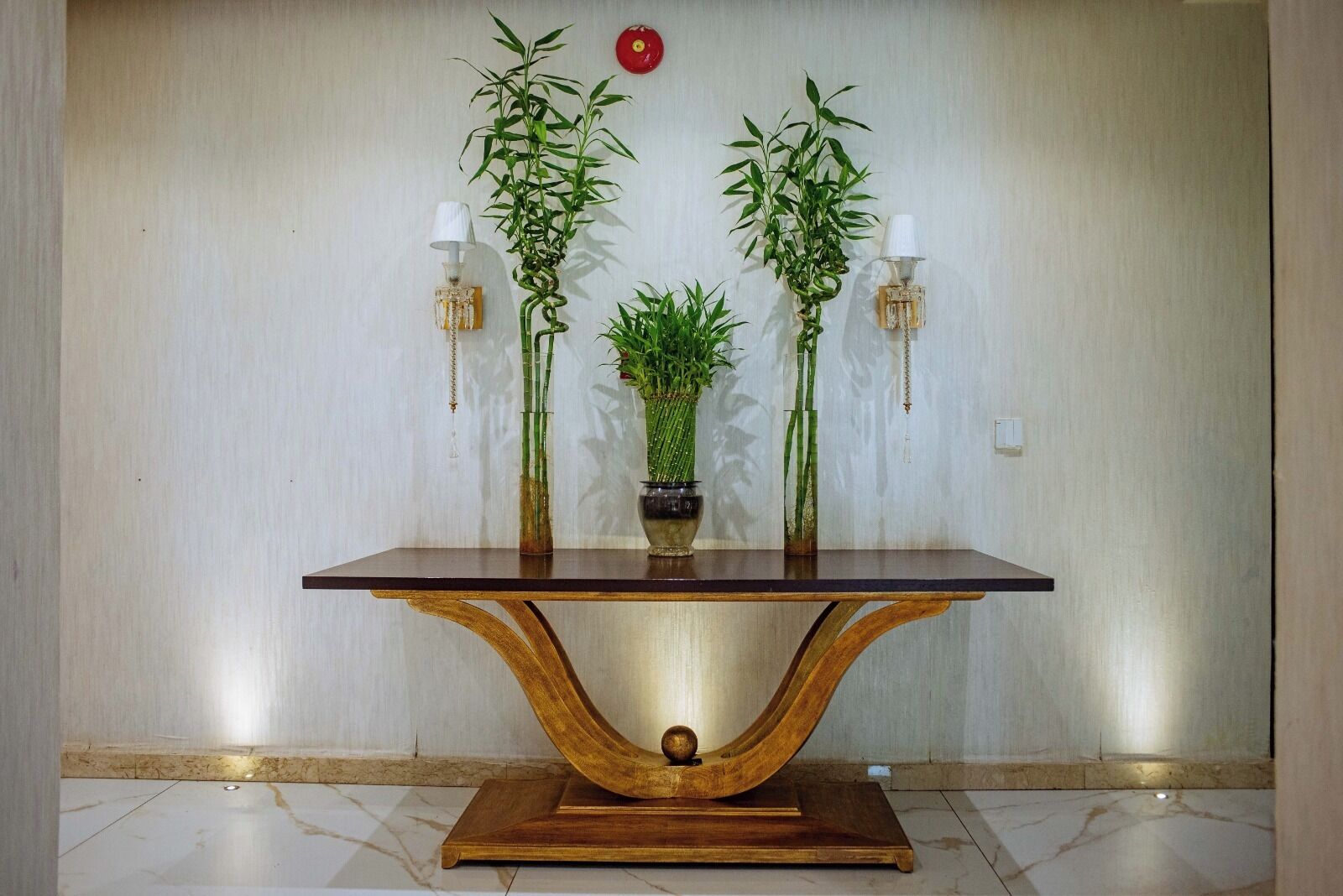 Una mesa de madera con cubierta de vidrio, acompañada por dos macetas y dos lámparas de pared.