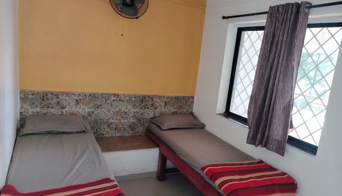 A room with two single beds, a small bench, a fan, and a window with curtains.
