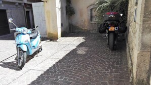 A narrow cobblestone street with a blue scooter parked on the left and a motorcycle on the right.