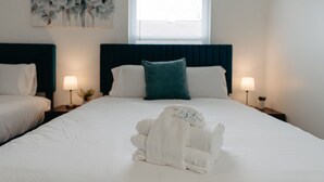 A neatly made bed with white linens and a folded towel in the center. There are two bedside tables with lamps, a window with blinds, and a wall-mounted painting. - Pondside Retreat 4BR w/ Hot Tub & Pool Table & BBQ (Hilliard)