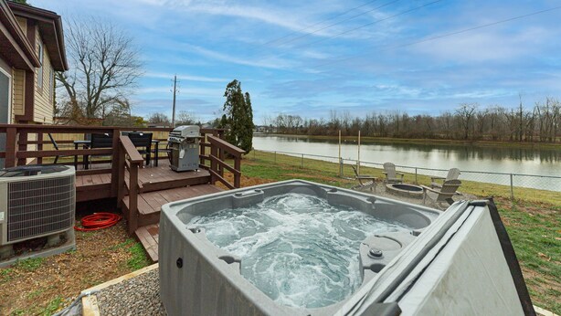 A hot tub on a deck with a view of a lake, outdoor furniture, and a grill. - Pondside Retreat 4BR w/ Hot Tub & Pool Table & BBQ (Hilliard)
