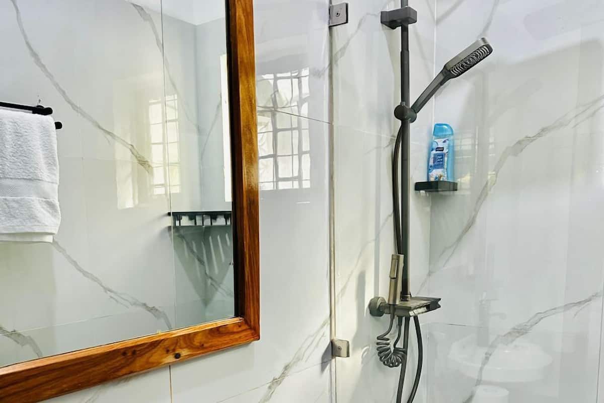 Casa de banho com soalho em mármore, espelho com moldura de madeira, lavatório branco, sanita e cabine de duche com divisórias de vidro, chuveiro manual e torneira manual.