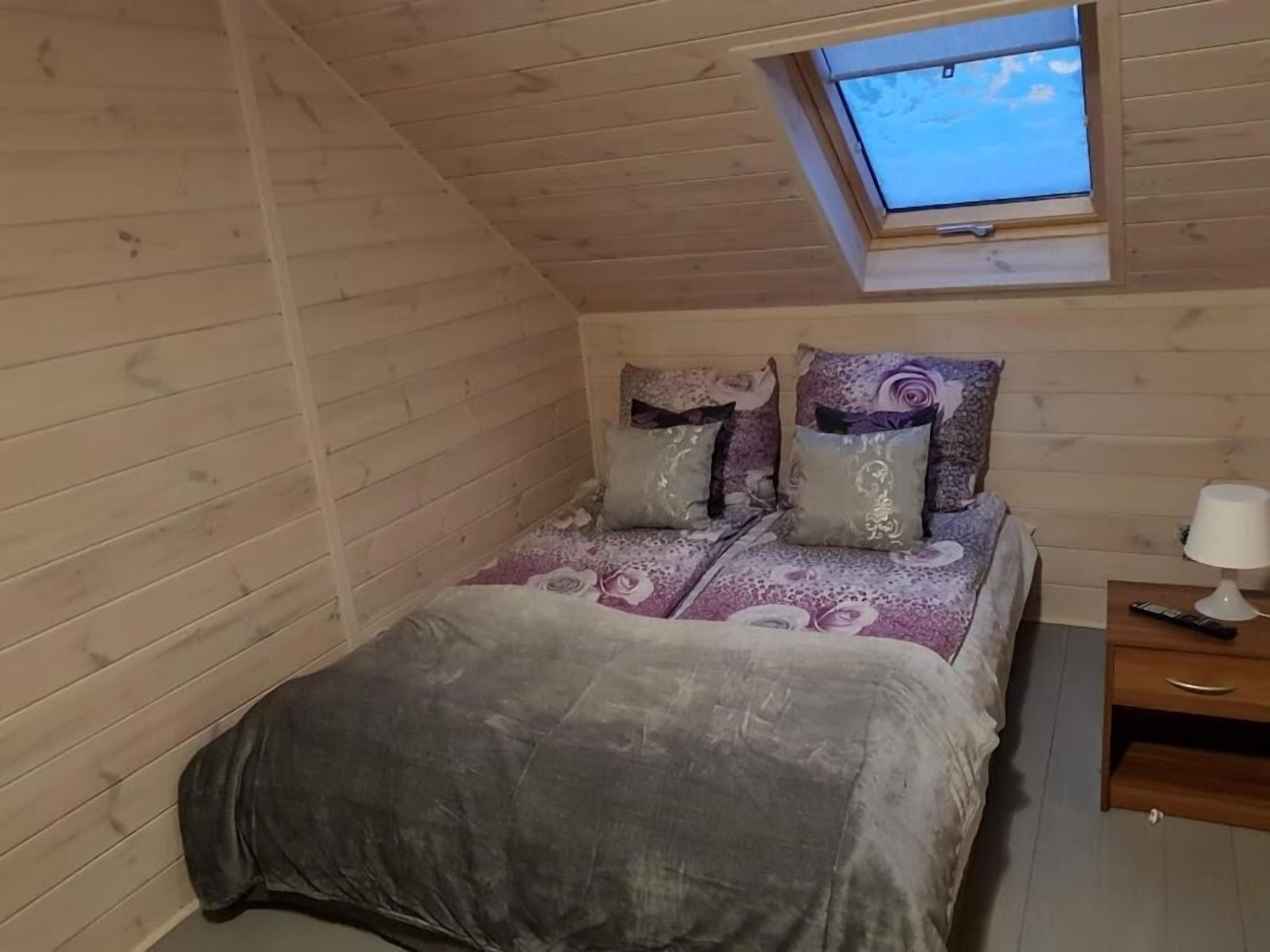 A small, cozy room with a sloped ceiling and a skylight. It features a bed with a patterned bedspread and pillows, a bedside table with a lamp, and a wooden nightstand.
