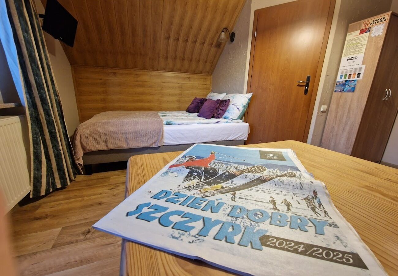 Um quarto pequeno e acolhedor com teto de madeira e uma cama com colcha branca e roxa. Um cartaz sobre a mesa diz "DEJEN DO BZYRZ 2024/2025".