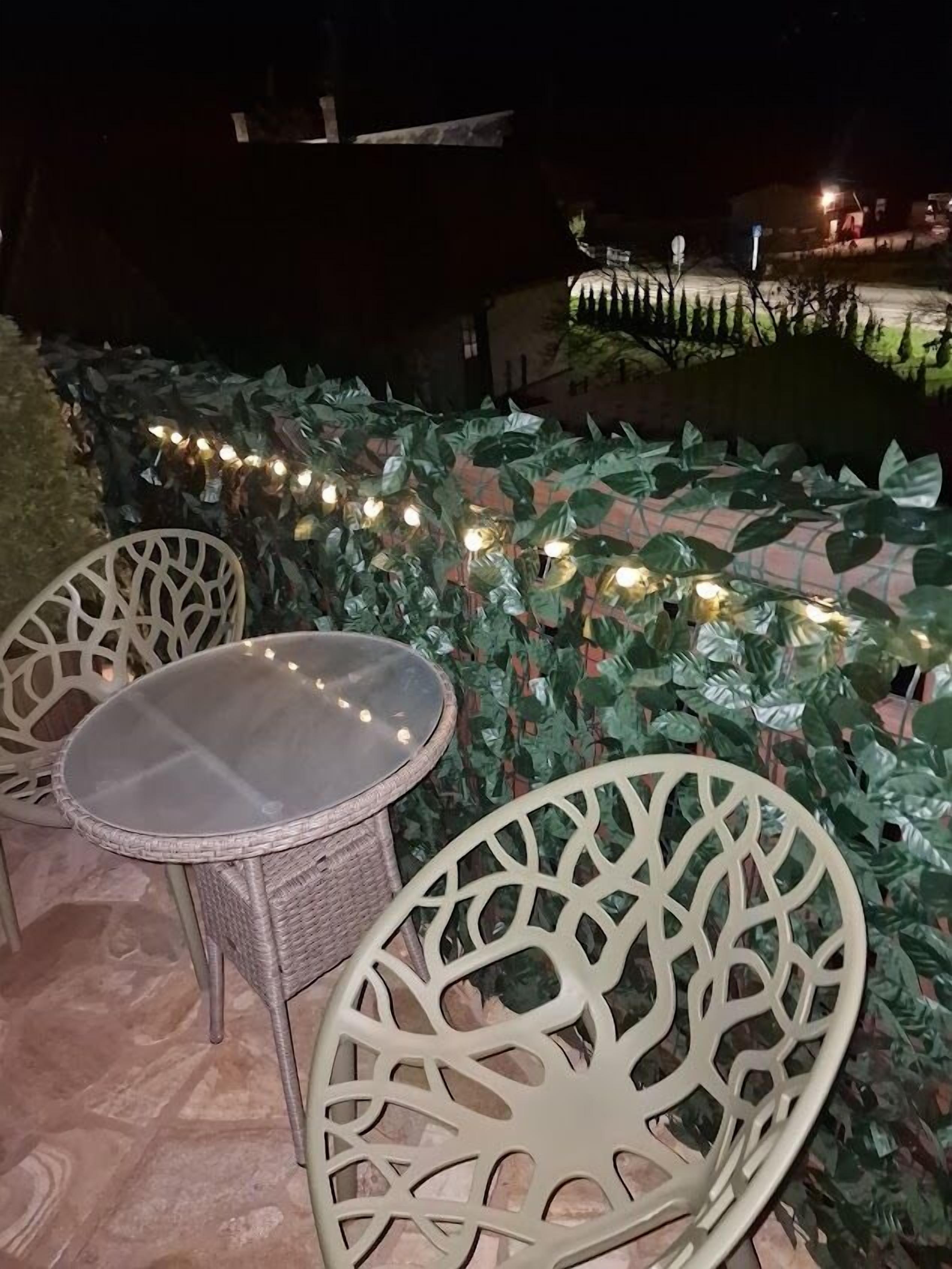 Un área de estar al aire libre con sillas de mimbre y una mesa, rodeada de abundante follaje y luces colgantes.