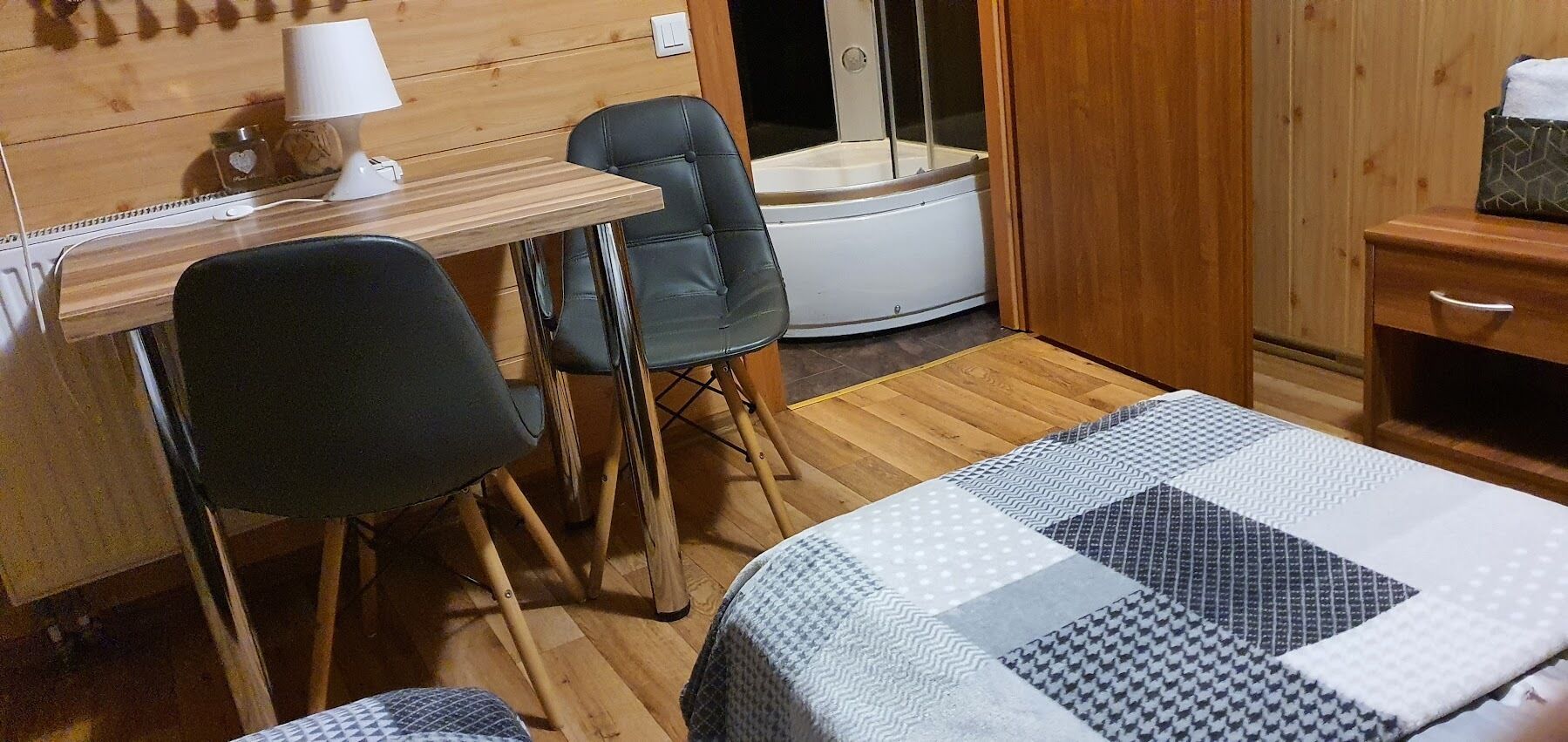 A small, cozy room with a wooden desk, two chairs, a bed with a patterned quilt, and a shower area.