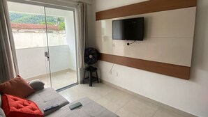 Ein modernes Wohnzimmer mit grauem Sofa, roten Kissen, einem flachbildfernseher an einer Holzvertäfelung, einem Standventilator und einer Glastür, die auf einen Balkon führt.