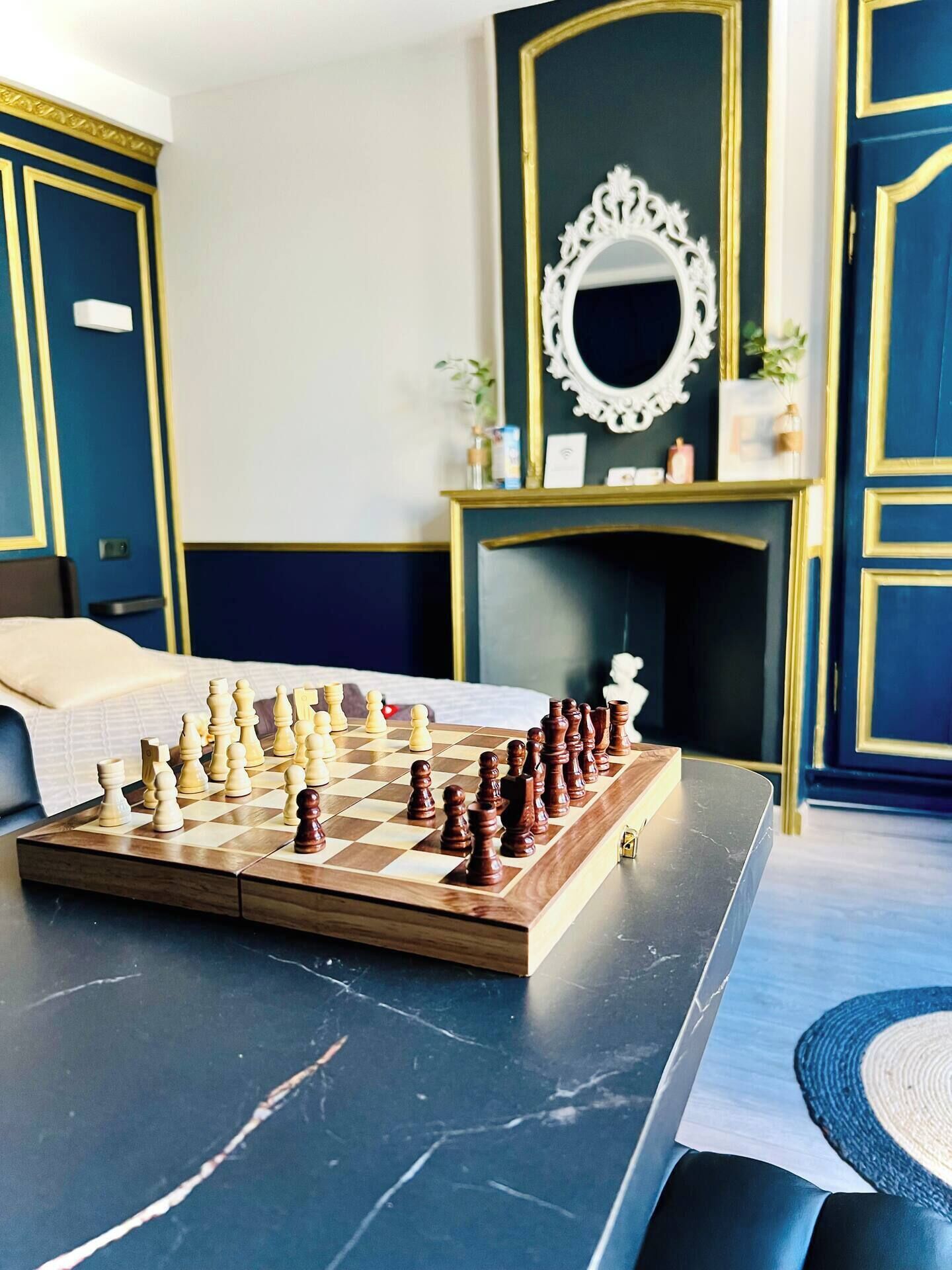 Ein Schachbrett auf einem Tisch in einem Raum mit blauen und goldenen Farbtönen, einem Kamin und einem Spiegel darüber.