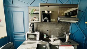 A compact kitchen with a microwave, coffee maker, and a small refrigerator. There is a black chair and a table with a black and white patterned tablecloth. The kitchen has a blue accent wall with geometric patterns. - Studio Royal (Saint-Omer)
