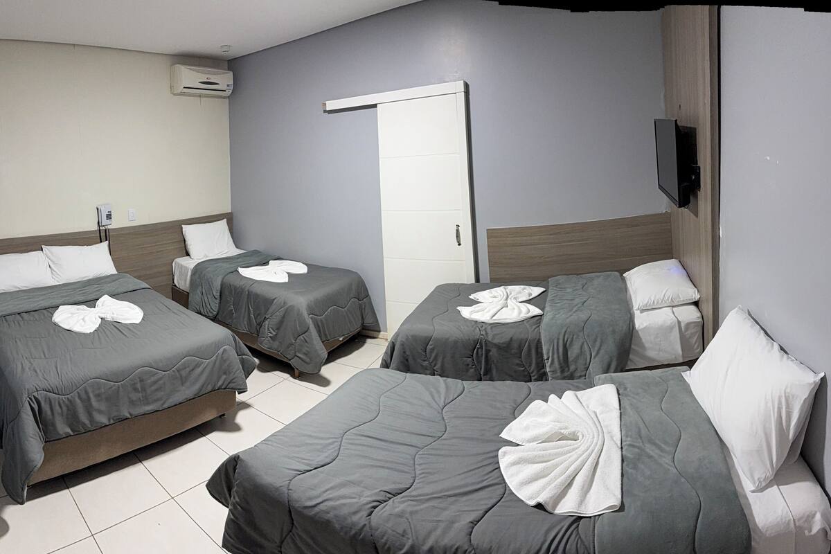 A hotel room with three beds, each with a towel and pillow. There is a door, a wall-mounted air conditioner, and a flat-screen TV.