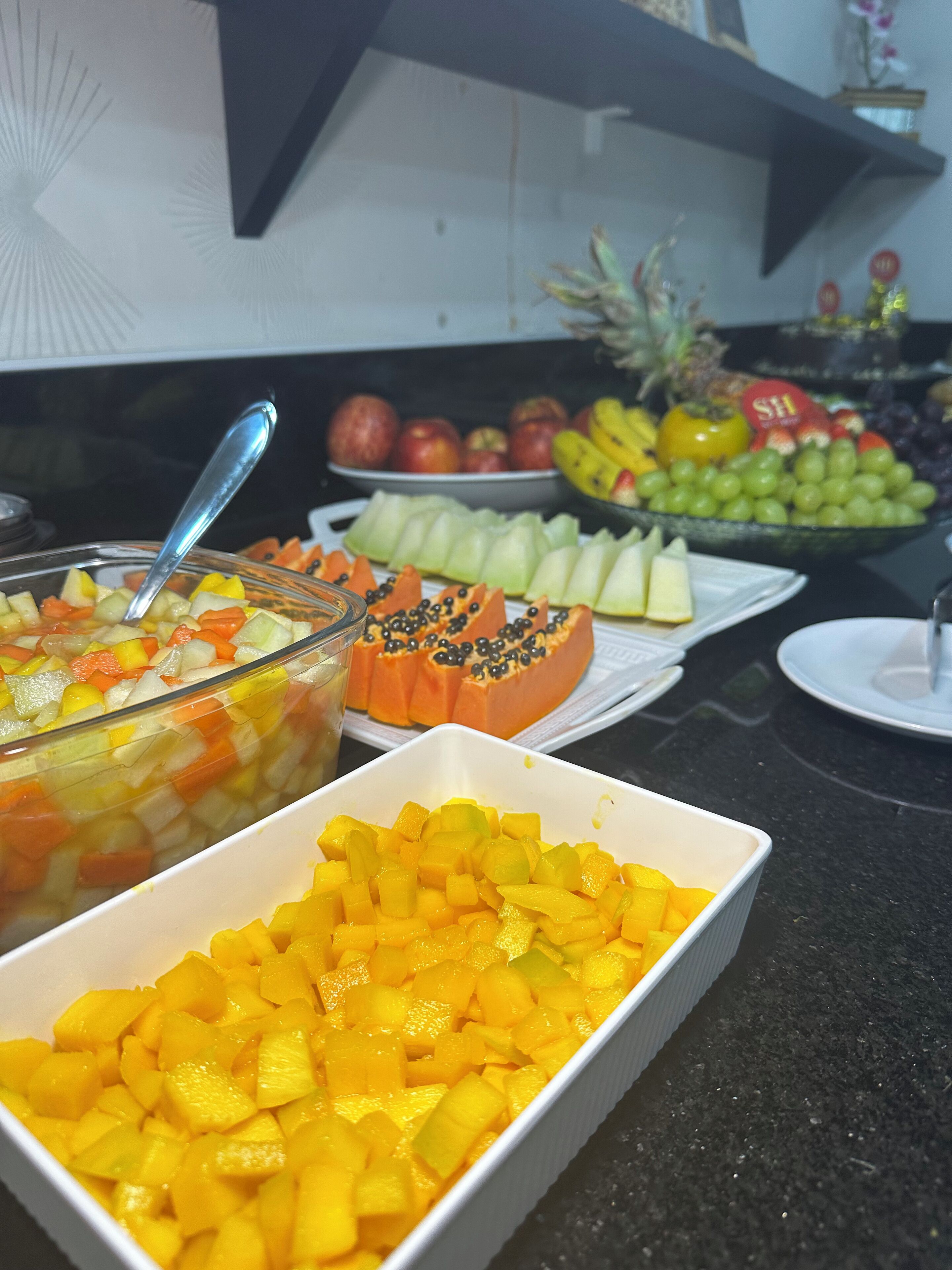 A buffet table with various fruits and vegetables, including a bowl of mixed fruit salad, a plate of sliced papaya, and a tray of diced mango.