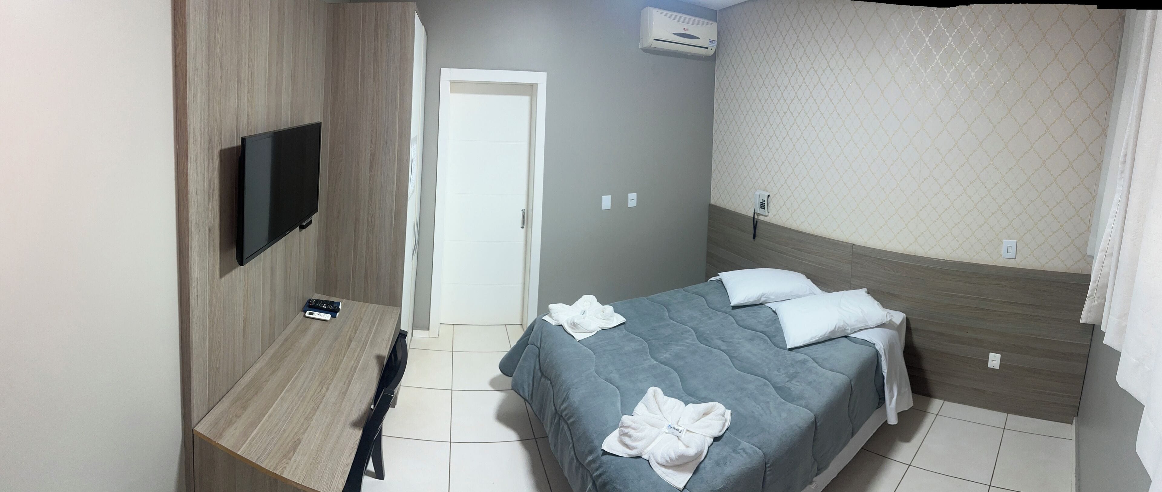 A hotel room with a bed, a TV mounted on the wall, a door, and an air conditioning unit.