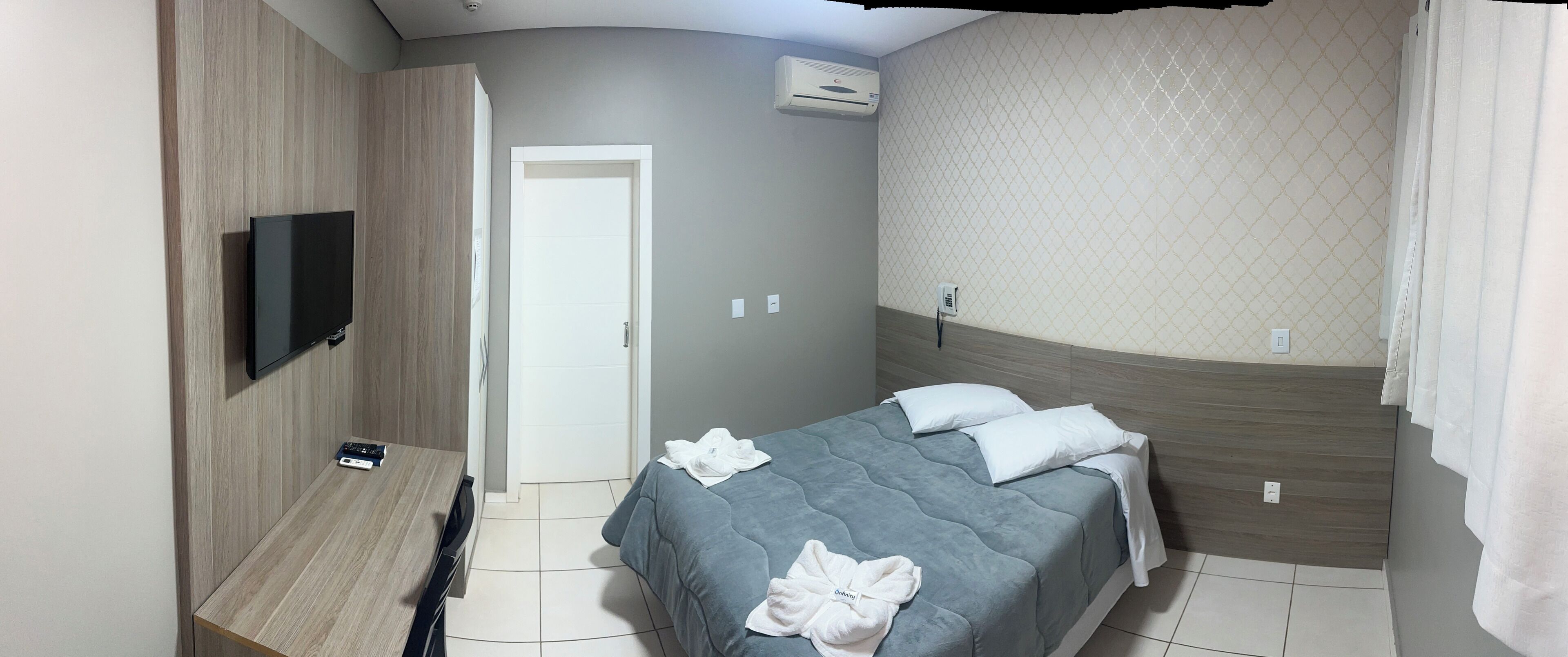 Habitación de hotel con cama, televisor montado en la pared, escritorio y mando a distancia, además de una puerta.