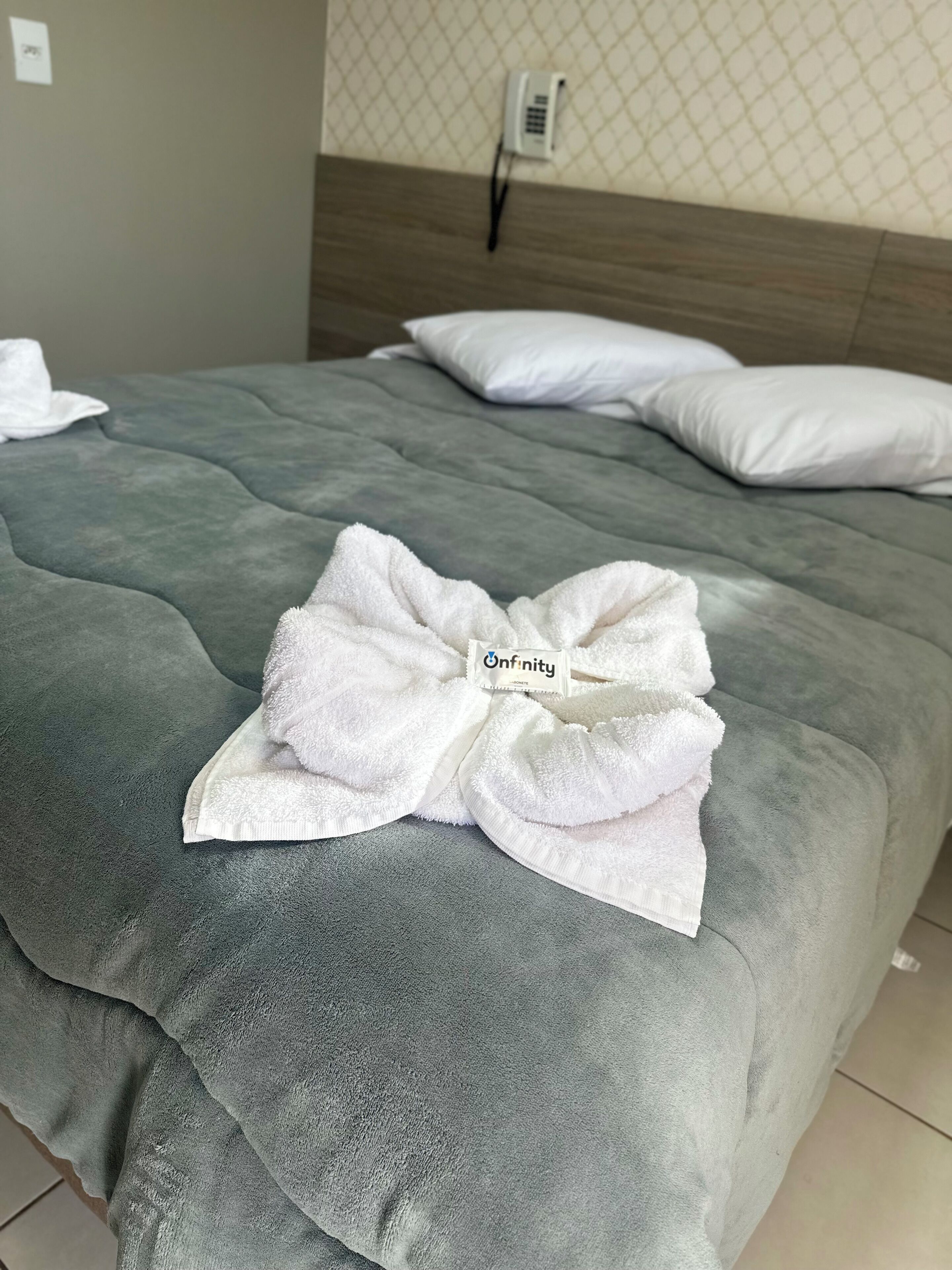 A neatly made bed with a grey duvet, a white pillow, and a white towel folded into a bow with a label reading "Infinity".