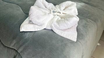 A neatly made bed with a grey duvet, a white pillow, and a white towel folded into a bow with a label reading "Infinity".