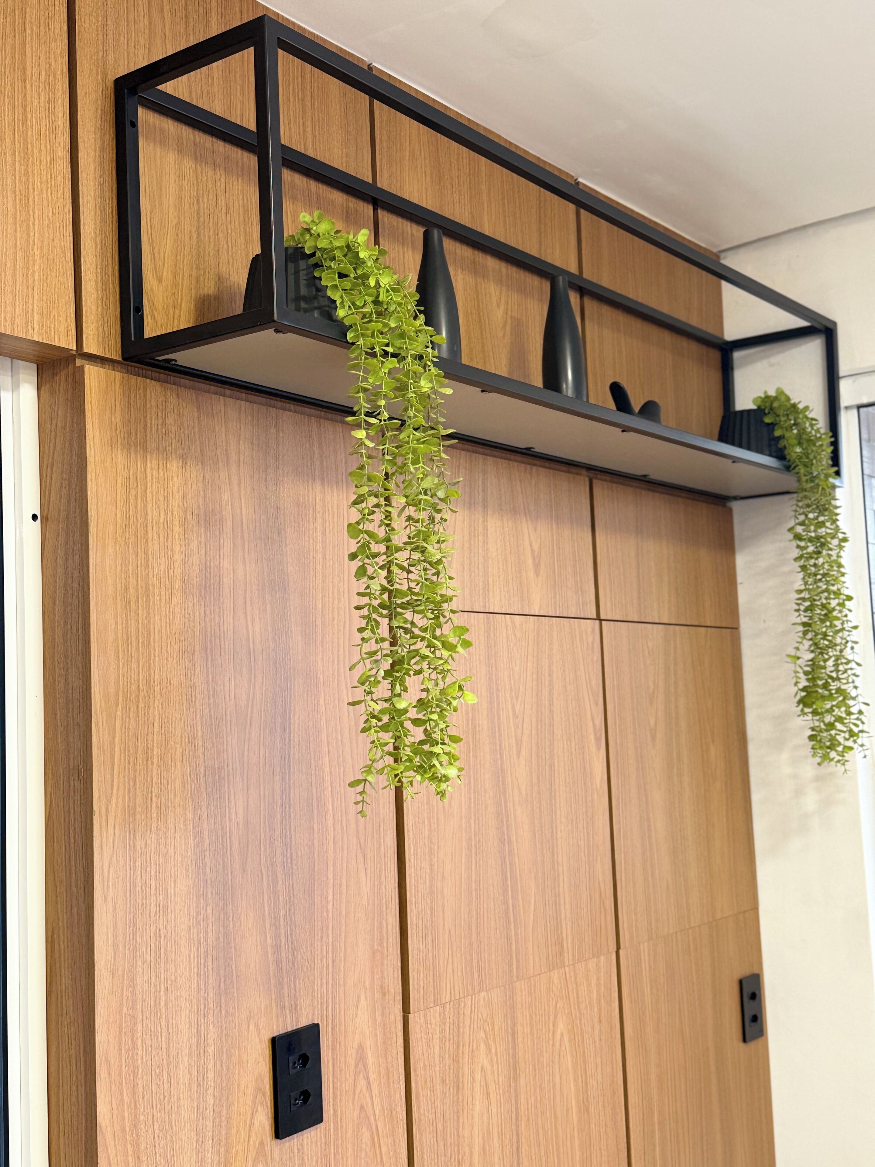 A wooden wall with a black metal shelf holding two black vases and a hanging green plant.