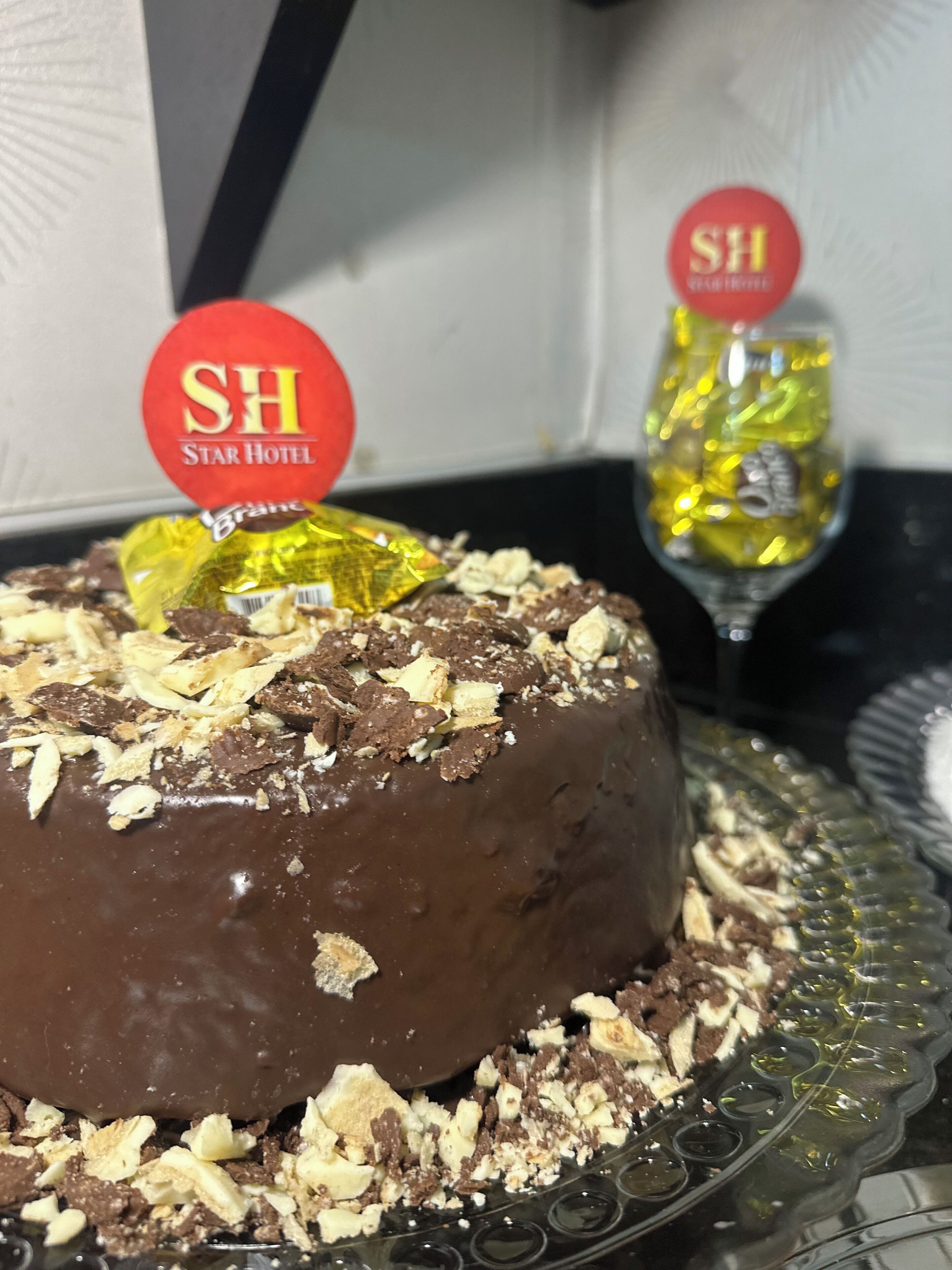 A chocolate cake with a Star Hotel logo on top, placed on a glass cake stand.