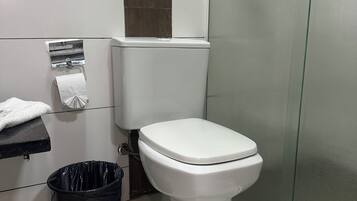 A bathroom with a toilet, a trash bin, a towel rack, and a glass shower enclosure.