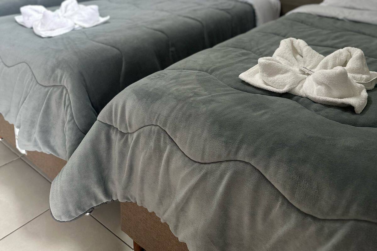 Two beds with grey bedding and white towels, a wall-mounted air conditioner, and a plain wall with a light switch.