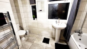 Une salle de bain avec des toilettes, un lavabo et une baignoire. On y trouve un porte-serviettes et une petite fenêtre avec une plante.