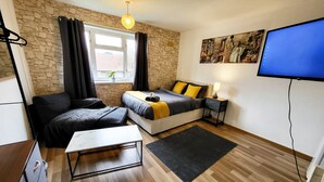 Une chambre moderne avec un mur en pierre, un lit recouvert d’une couverture jaune, un bureau, une chaise, un téléviseur et une fenêtre avec des rideaux.