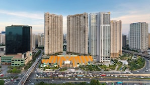 A high-rise residential building complex with multiple apartment buildings, a central commercial building with a distinctive orange facade, and a busy multi-lane road with moderate traffic. - TIL Vinhomes D'capitale Elegance Suites (Hanoi)