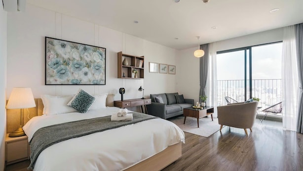 A modern hotel room with a large bed, a sofa, a chair, and a balcony with a view. - TIL Vinhomes D'capitale Elegance Suites (Hanoi)
