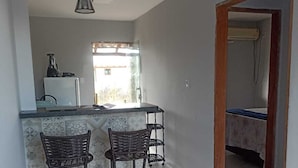 A modern kitchen with a white refrigerator, a blender, and a window. There are two wicker bar stools and a beige sofa in the living area. - FLAT SHELDOM (Maraú)