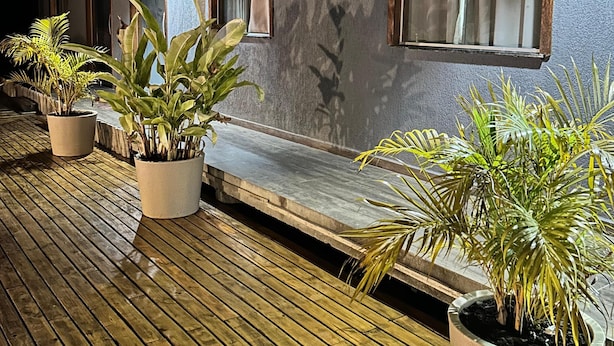 A wooden deck with potted plants, a covered seating area, and a building with a sloped roof and large windows. - FLAT SHELDOM (Maraú)