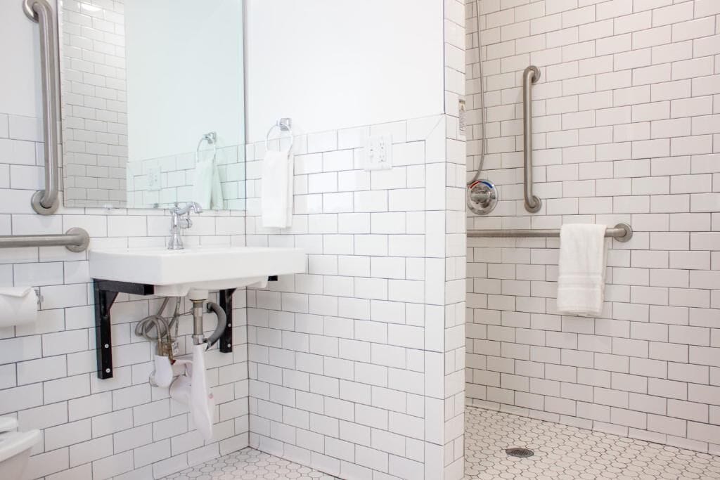 Une salle de bain avec des murs recouverts de carreaux blancs, un lavabo avec un miroir et un espace douche équipé de barres d’appui.