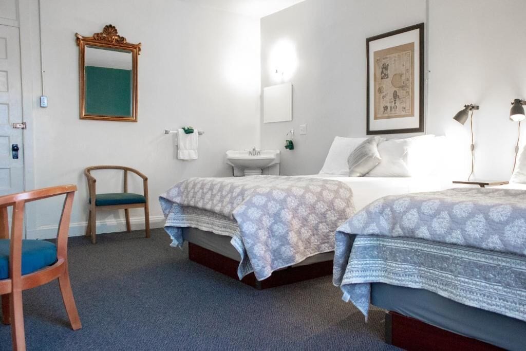 A hotel room with two beds, a wooden chair, a sink, and a mirror.
