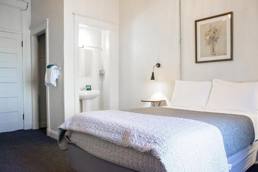 A hotel room with a bed, a bedside table with a lamp, a framed picture on the wall, and a bathroom visible through an open door.