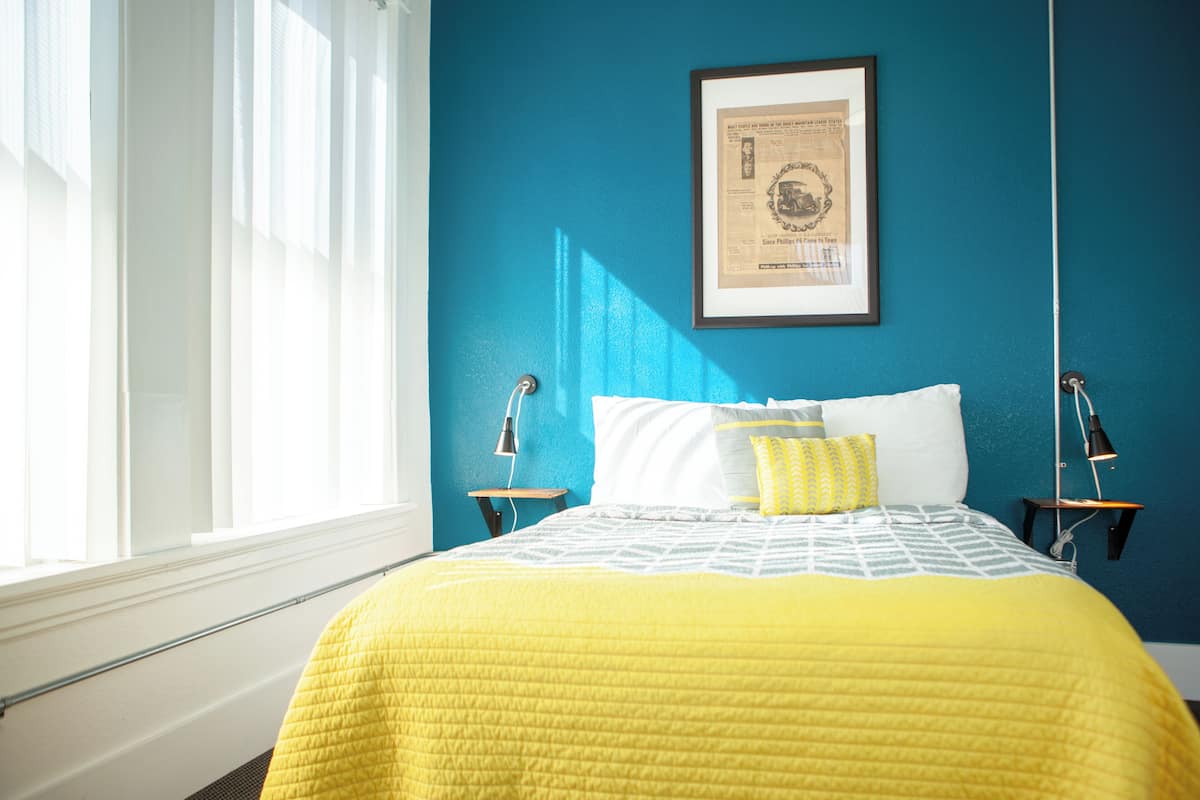 A bedroom with a bed, two bedside tables, a framed certificate on the wall, and a window with blinds.