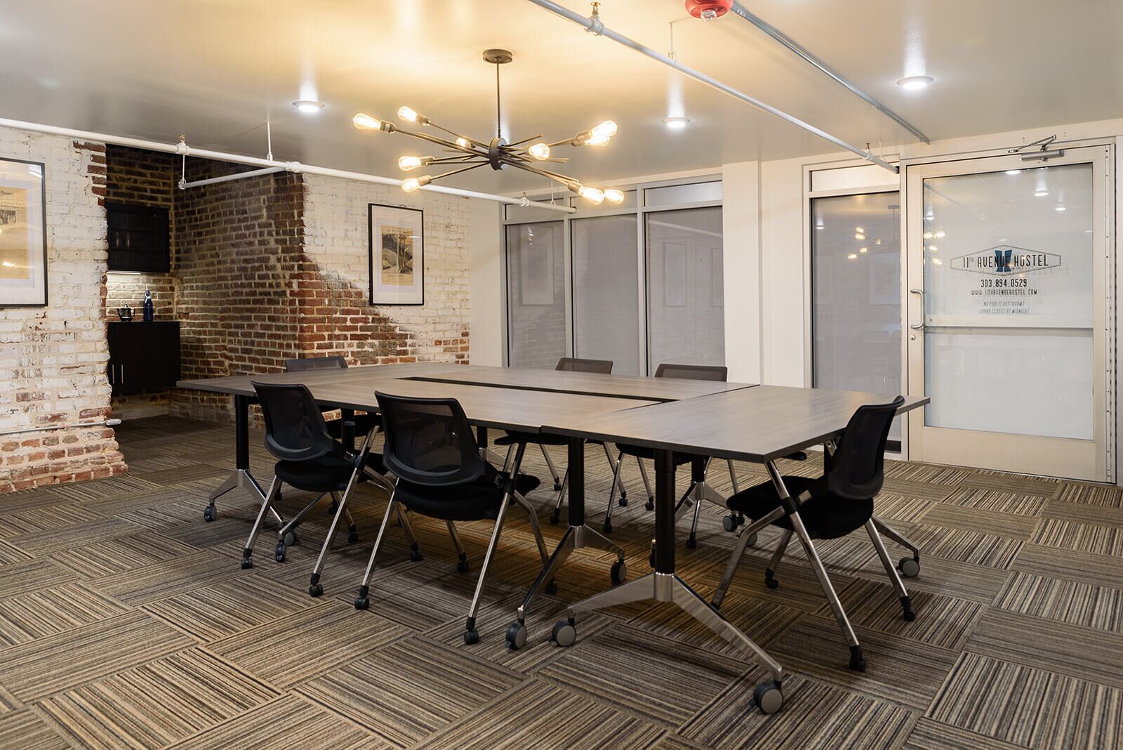 Uma sala de conferências com uma mesa comprida, cadeiras pretas, parede de tijolos e luminária no teto.