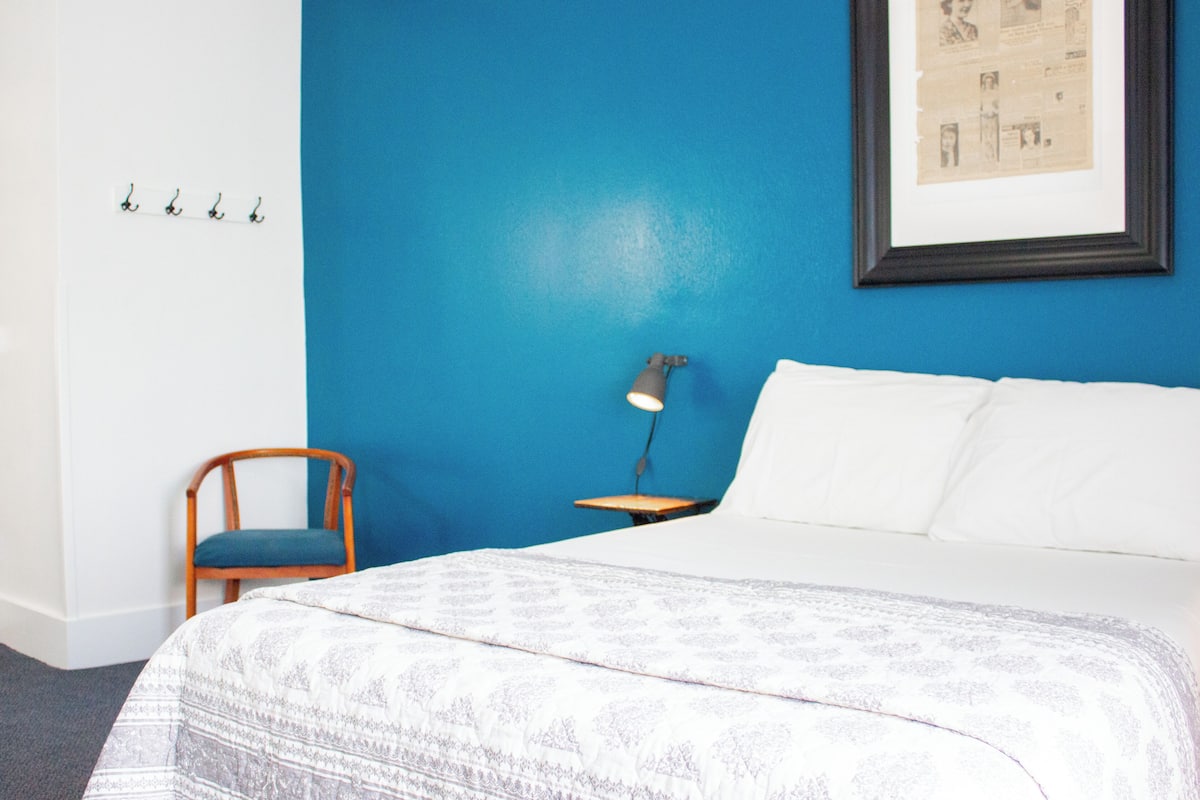 A bedroom with a bed, a chair, a desk with a lamp, and a framed picture on the wall.