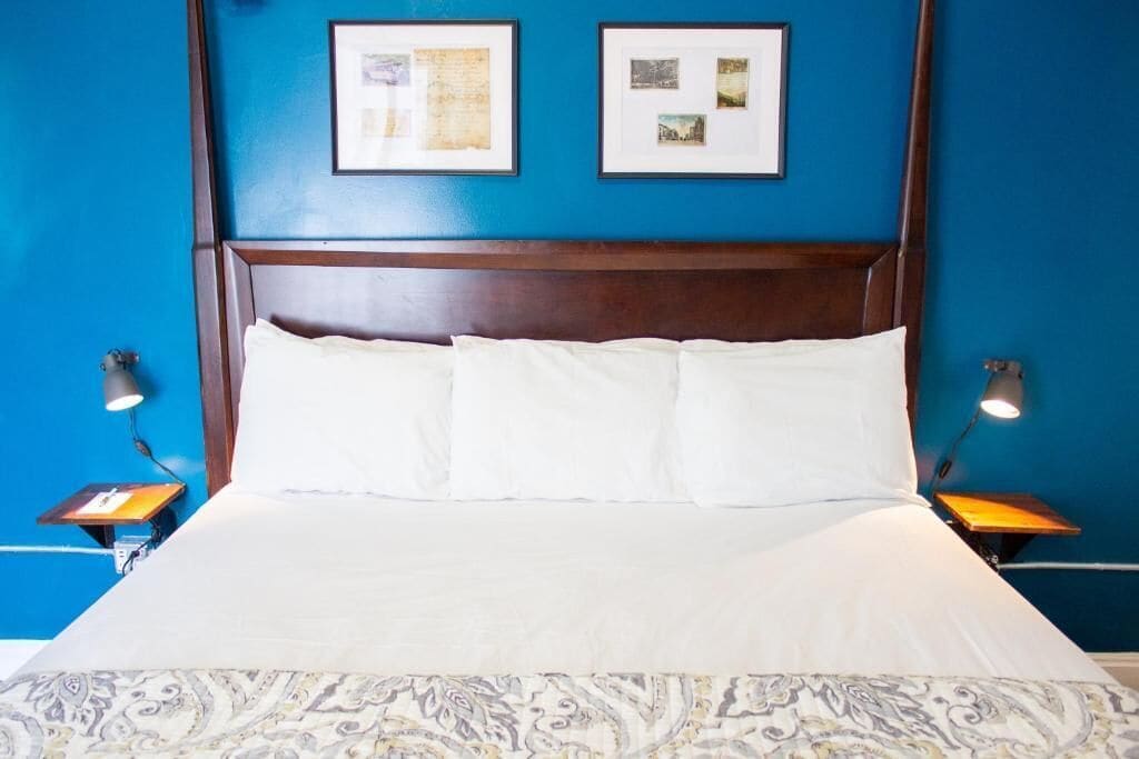 A bed with white linens and a patterned bedspread, flanked by two bedside tables with lamps, in a room with blue walls and framed pictures above the bed.