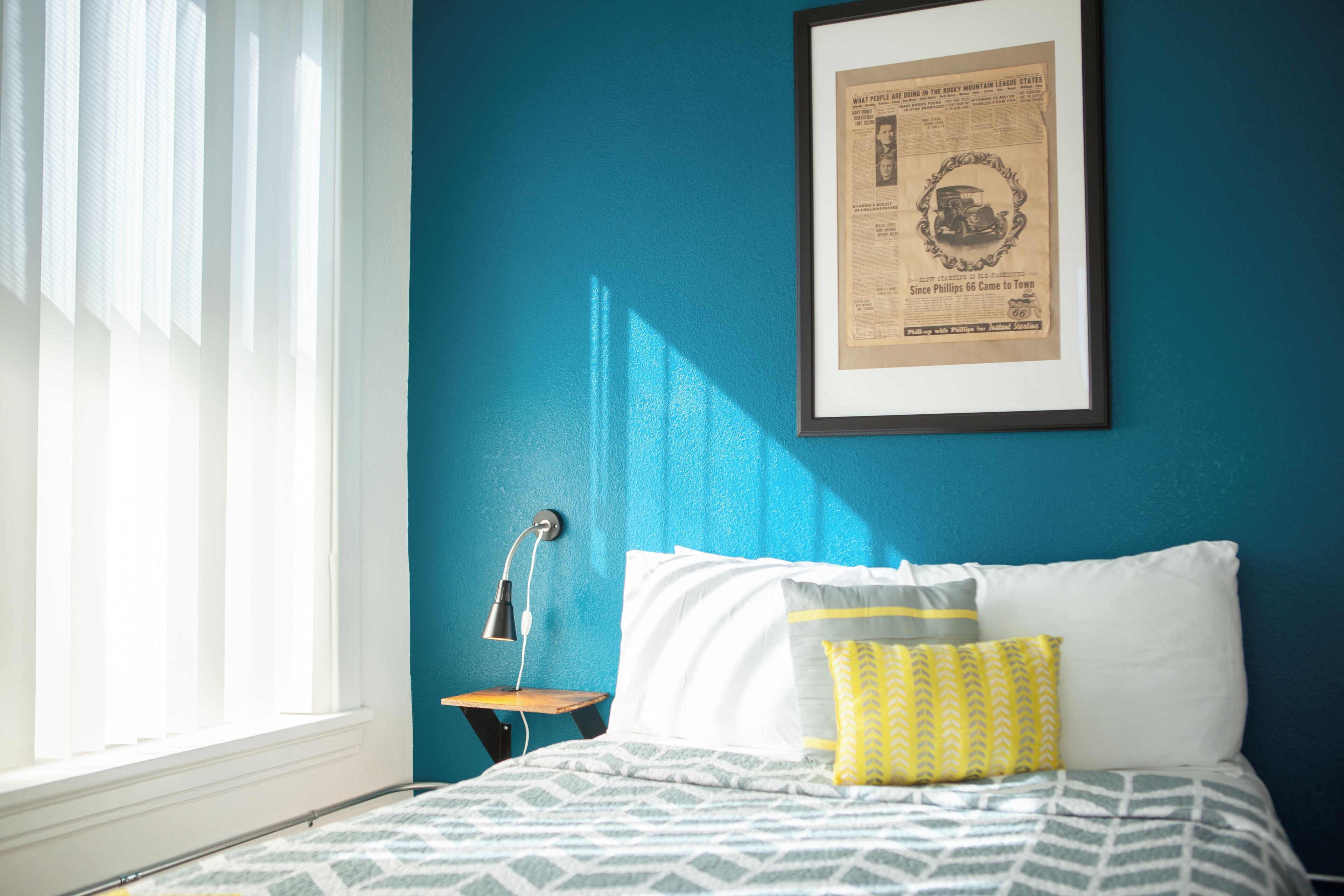 A bedroom with a bed, pillows, a framed picture on the wall, a nightstand, and a window with blinds.