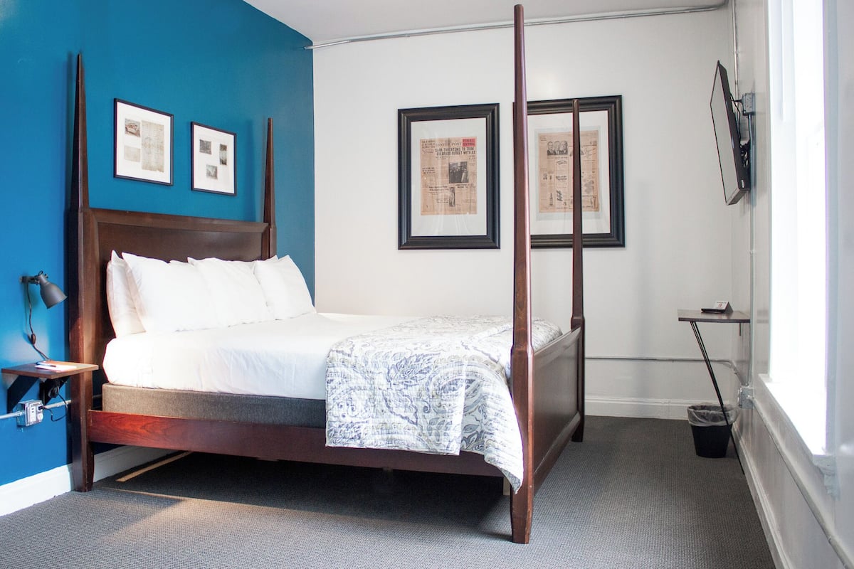 A four-poster bed with white linens, a small bedside table with a lamp, and a framed newspaper on the wall.