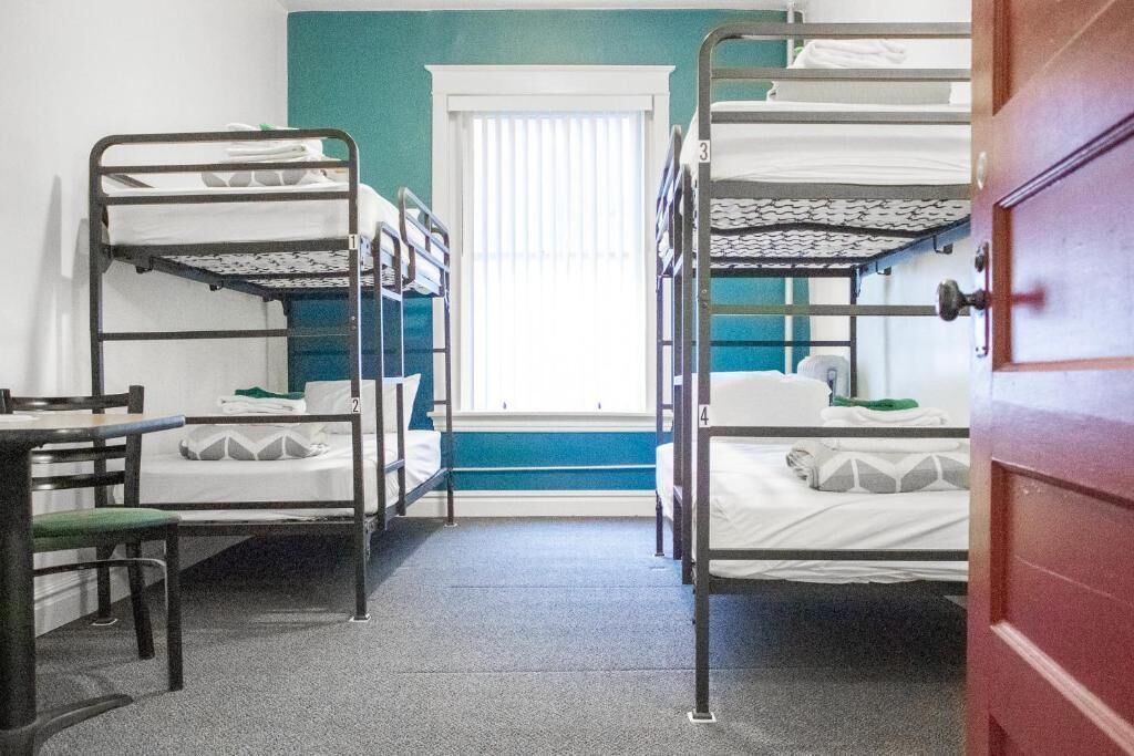A dormitory room with bunk beds, a window with blinds, a desk, and a chair.