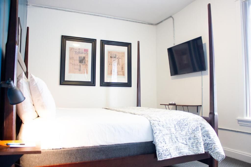 A bedroom with a bed, a desk with a lamp, two framed pictures on the wall, and a TV mounted on the wall.