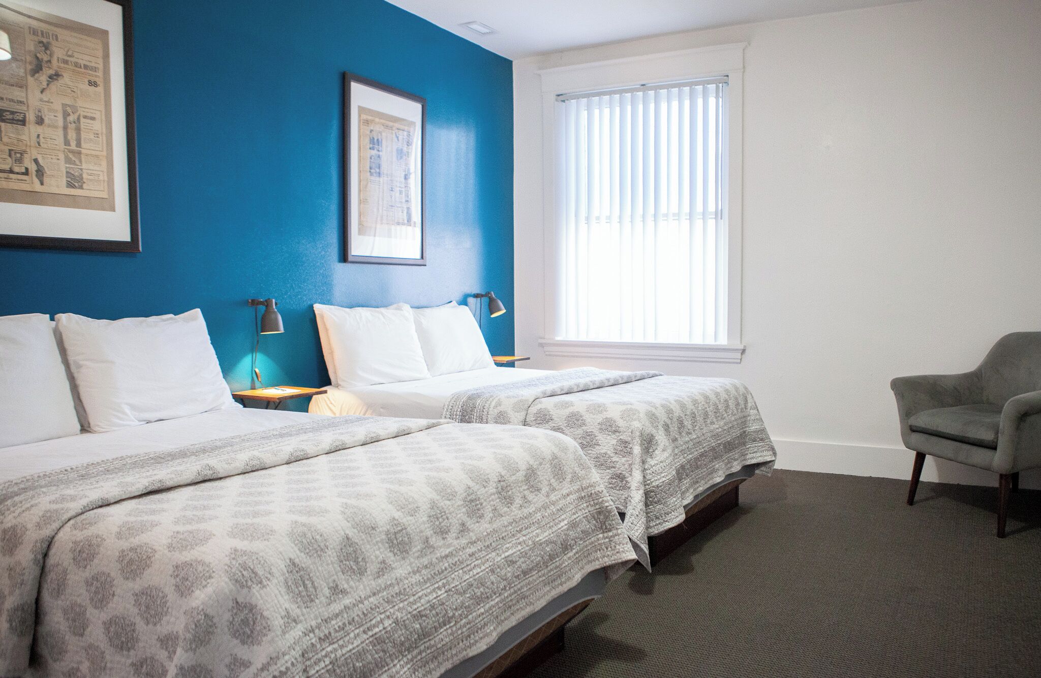 A hotel room with two beds, a chair, a window with blinds, and a framed picture on the wall.