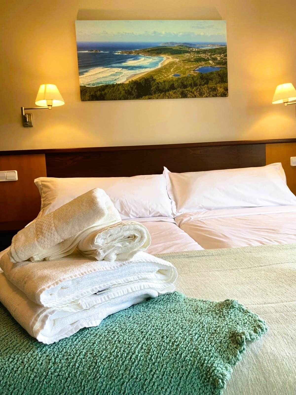 Un lit bien fait, avec des draps blancs, un plaid vert et, au-dessus, un tableau représentant un paysage marin.