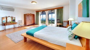 A spacious bedroom with a large bed, a wooden bench, a desk, and a view through the windows.