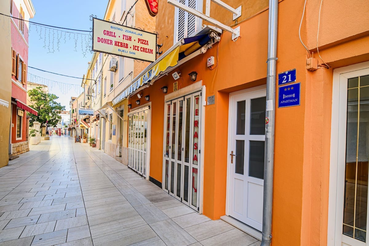 Eine schmale Straße mit einem Restaurant namens "BISTRO - ZDRAVLJAK", das Grillgerichte, Fisch, Pizza, Döner Kebab und Eis anbietet.