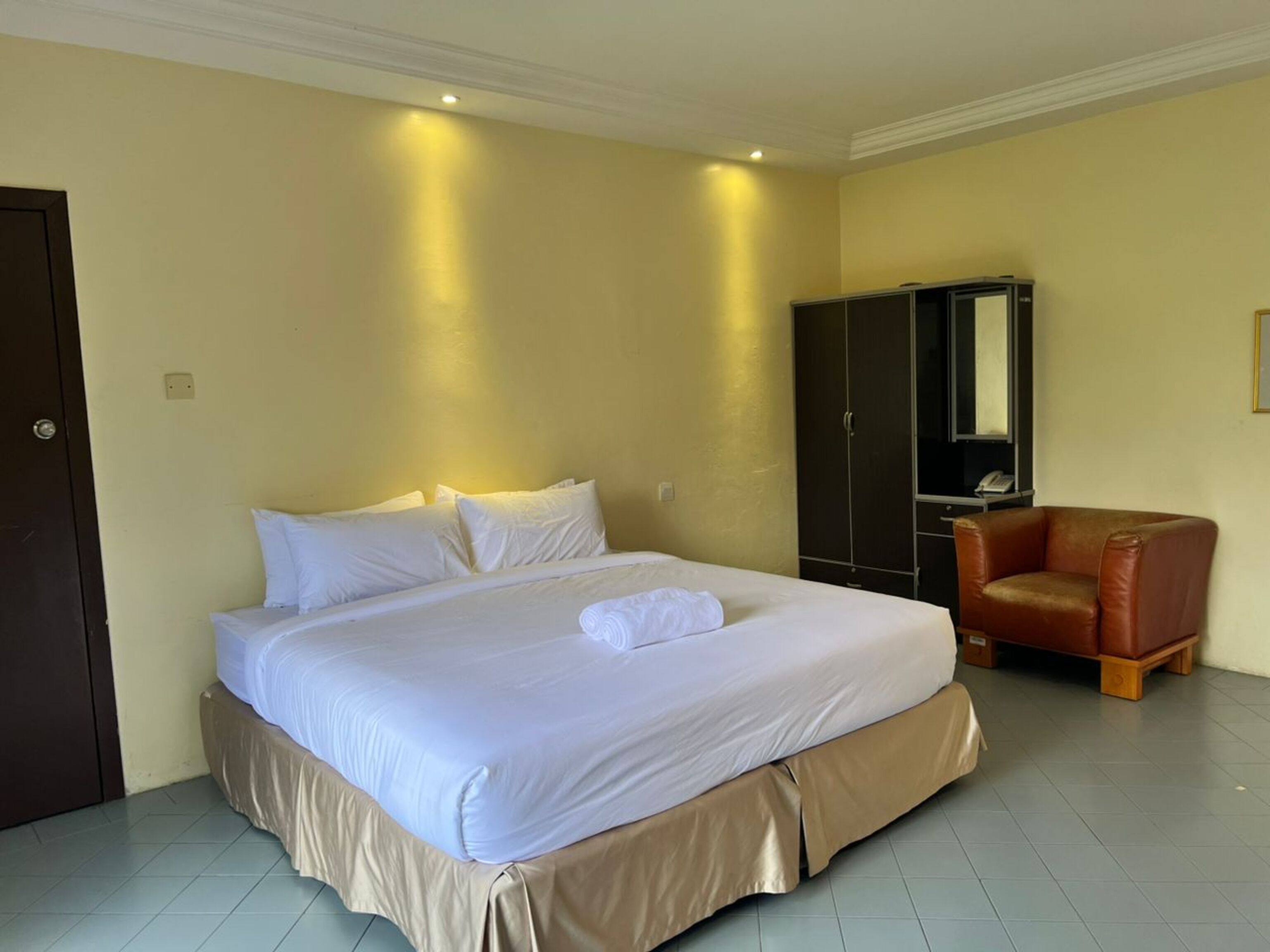A hotel room with a large bed, a brown leather chair, a wardrobe, and a mirror.